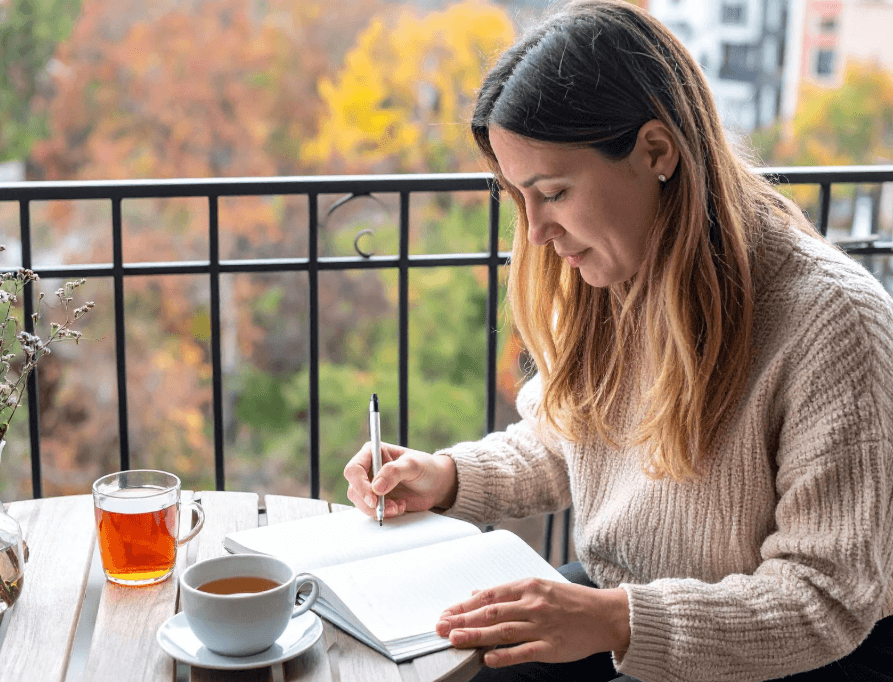 Staying healthy and happy during the cold: Woman writing with tea, cozy sweater, outdoor balcony autumn scene. Cold weather wellness.