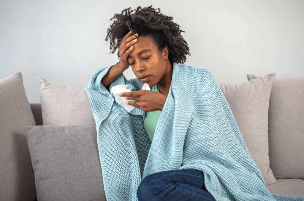 Woman with winter illness, wrapped in blanket, holding tissue. Preparation for cold and flu season during winter surge.