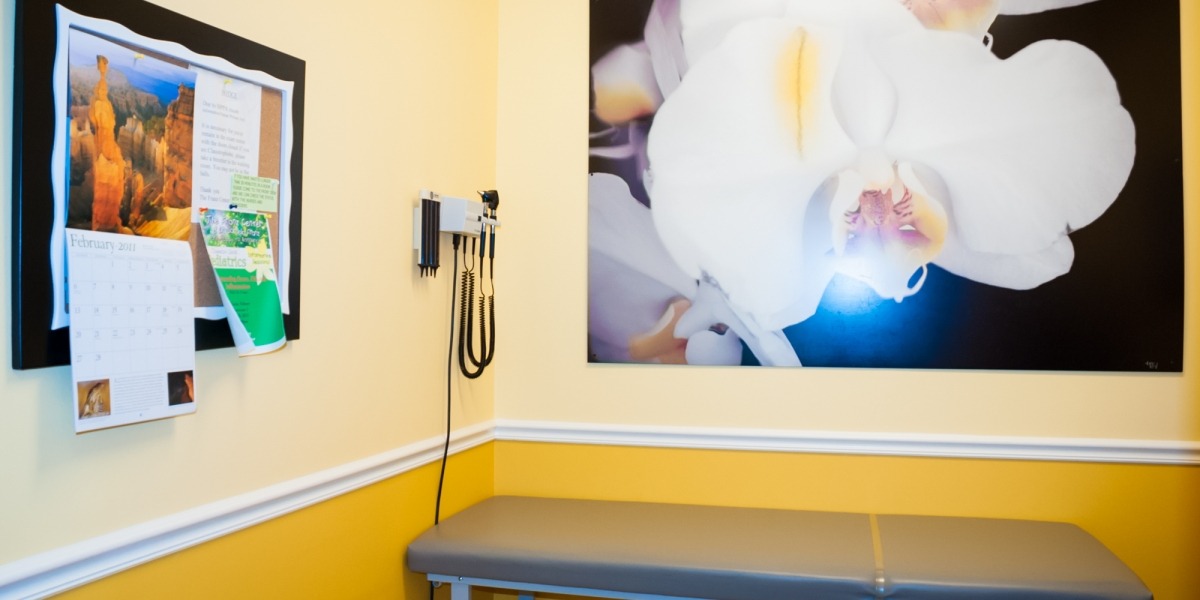 Yellow-themed doctor's office with examination table, orchid art, and a calendar offering a welcoming and professional About space.